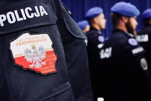 Grupa policjantów. Na pierwszym planie naszywka na ramieniu munduru w barwach biało-czerwonych w kształcie mapy Polski. Na konturze Polski godło. Powyżej naszywki napis: Policja.
