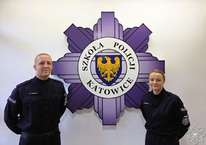 Policjant i policjantka pozują do zdjęcia na tle ściany z logo szkoły Policji w Katowicach.
