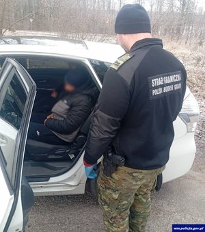 Funkcjonariusz straży granicznej stoi przy samochodzie, w którym siedzą dwaj nielegalni imigranci.