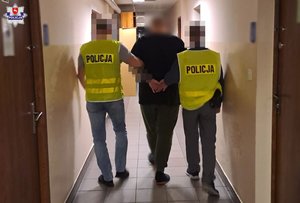 Dwóch policjantów w żółtych kamizelkach prowadzi zatrzymanego mężczyznę korytarzem.