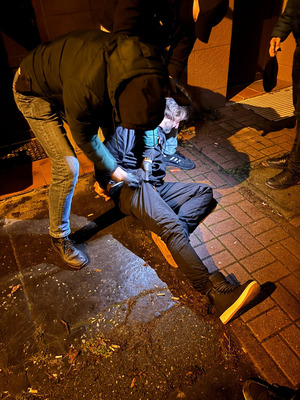 Policjant przeszukuje zatrzymanego mężczyznę leżącego na ziemi.