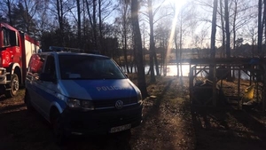 Radiowóz policyjny i wóz strażacki stoją między drzewami, w głębi zbiornik wodny.