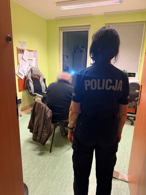 Zatrzymany siedzi na krześle w pomieszczeniu. W drzwiach do pomieszczenia stoi policjantka odwrócona tyłem.