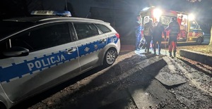 Na pierwszym planie radiowóz policyjny. W głębi policjanci prowadzą mężczyznę do karetki pogotowia.