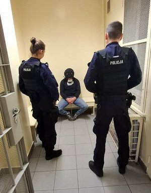 Zatrzymany siedzi na ławce skuty w kajdanki. Naprzeciwko niego stoi policjant i policjantka.