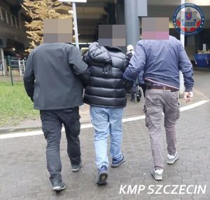 Dwaj policjanci prowadzą mężczyznę zakutego w kajdanki w kierunku budynku.