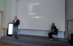 Mężczyzna stoi podczas wystąpienia w trakcie konferencji na tle ekranu, na którym wyświetlana jest prezentacja. Po prawej stronie na krześle siedzi kobieta.