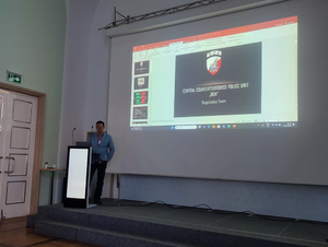 Mężczyzna stoi przy mównicy podczas wystąpienia w trakcie konferencji na tle ekranu, na którym wyświetlana jest prezentacja.