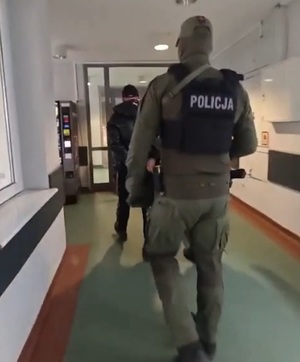 Idący korytarzem, widziany od tyłu funkcjonariusz, a przed nim zatrzymany mężczyzna w kajdankach.