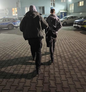 Policjant prowadzi przez parking w kierunku budynku komendy Policji zatrzymanego mężczyznę zakutego w kajdanki.