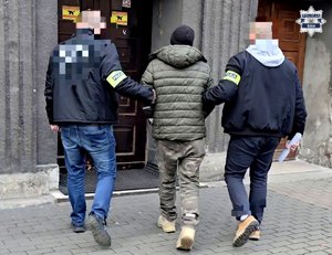 Zatrzymany w kajdankach zespolonych prowadzony przez dwóch nieumundurowanych policjantów, przed wejściem do prokuratury.