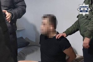 Dwaj policjanci z zatrzymanym. Jeden z policjantów trzyma dłoń na ramieniu zatrzymanego, który siedzi na łóżku.