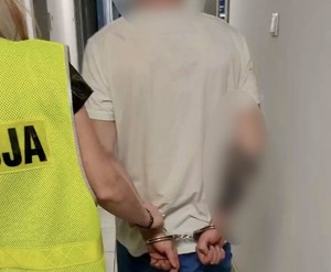 Zatrzymany w kajdankach założonych na ręce trzymane z tyłu. Obok niego policjant trzymający go za ramię.