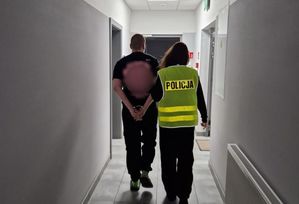 Policjantka w żółtej kamizelce z napisem Policja na plecach prowadzi zatrzymanego.