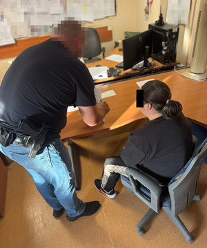 zatrzymana siedzi przy biurku. obok stoi policjant.