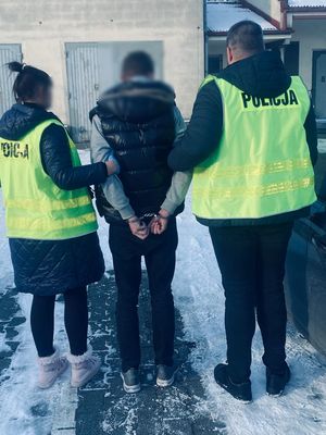 Dwoje policjantów w żółtych kamizelkach z napisem Policja na plecach, prowadzi zatrzymanego.