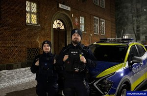 policjant i policjantka pozują do zdjęcia. stoją obok radiowozu.