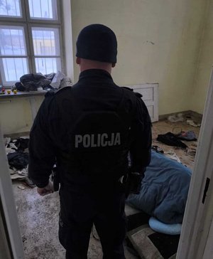 Wnętrze pustostanu, Policjant stoi obok leżącego pod materiałem na podłodze mężczyzny.