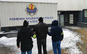Dwaj funkcjonariusze prowadzą zatrzymanego mężczyznę w kierunku budynku Komendy Wojewódzkiej Policji w Łodzi.