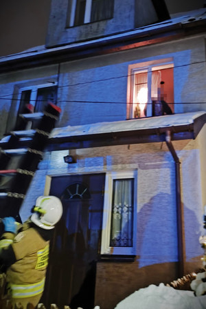 Strażak przystawia drabinę do ściany budynku. Przez okno na piętrze wygląda mężczyzna.