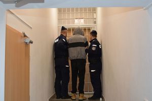 Dwaj policjanci prowadza zatrzymanego mężczyznę zakutego w kajdanki.