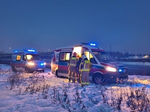 Dwie karetki pogotowia stoją jedna za drugą na brzegu rzeki. Przy jednej z nich stoją trzej strażacy.