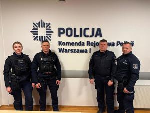 Trzej policjanci i policjantka pozują do zdjęcia na tle ściany z napisem: Policja Komenda Rejonowa Policji Warszawa I.