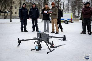 Na pierwszym planie dron, w tle policjanci, obok nich stoi kobieta, która pokazuje kartkę operatorowi drona.