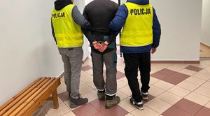 Dwaj policjanci w żółtych kamizelkach z napisem Policja na plecach prowadzą zatrzymanego.