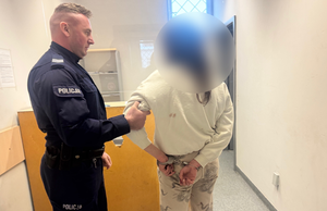Policjant trzyma zatrzymaną kobietę za ramię. Kobieta ma założone kajdanki na ręce trzymane z tyłu.