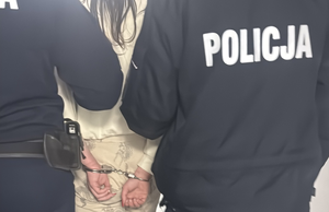 Ręce zatrzymanej kobiety zakute w kajdanki. Kobieta jest prowadzona przez dwóch policjantów.