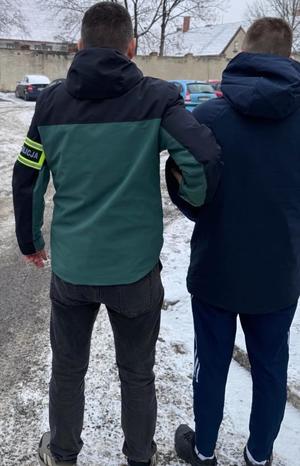 Funkcjonariusz trzyma za ramę zatrzymanego w kajdankach.