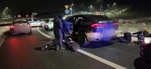 Policjant przy samochodzie i wokół zatrzymane osoby siedzące na ziemi.