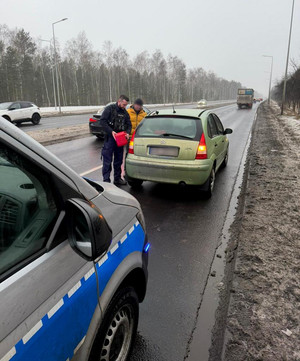 Policjant nalewa paliwo do baku stojącego auta. Z boku widać przejeżdżające pojazdy.