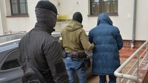 Dwaj funkcjonariusze prowadzą zatrzymaną osobę  w kierunku budynku.