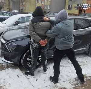 Policjant trzyma zatrzymanego mężczyznę zakutego w kajdanki przy samochodzie.