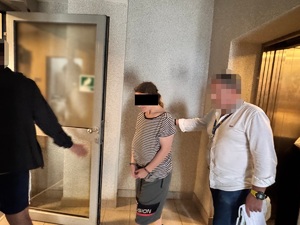 Zatrzymana kobieta prowadzona przez dwóch funkcjonariuszy korytarzem.