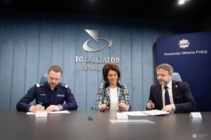 Komendant Główny Policji siedzi obok kobiety i mężczyzny z Totalizatora Sportowego. Komendant Główny Policji podpisuje dokumenty.