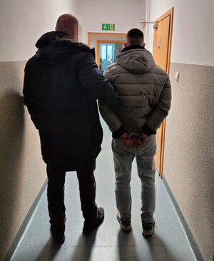 Funkcjonariusz trzyma zatrzymanego mężczyznę zakutego w kajdanki.