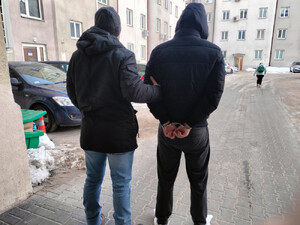 Funkcjonariusz trzyma zatrzymanego mężczyznę zakutego w kajdanki.