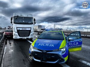 Na pierwszym planie radiowóz policyjny stojący przed samochodem osobowym wciśniętym między barierą a samochodem ciężarowym na autostradzie.