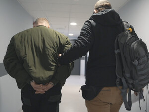 Mężczyzna zakuty w kajdanki prowadzony przez funkcjonariusza.