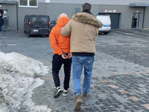 Mężczyzna zakuty w kajdanki prowadzony przez funkcjonariusza.