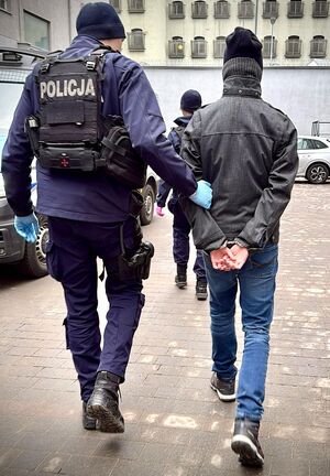 Funkcjonariusz prowadzi zatrzymanego mężczyznę zakutego w kajdanki.