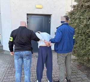 Dwaj funkcjonariusze trzymają zatrzymanego mężczyznę zakutego w kajdanki.