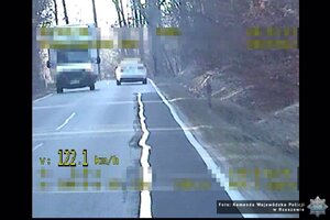 Widoczny samochód jadący przed radiowozem, z lewej strony widać przejeżdżające z przeciwka auta. W dolnym lewym rogu pojawia się żółty napis 122.1 km/h.