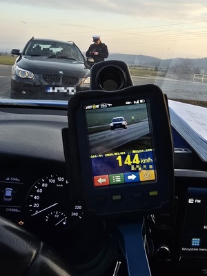 Widziany przez szybę radiowozu policjant przeprowadzający kontrolę drogową. Na pierwszym planie we wnętrzu radiowozu trzymany w dłoni prędkościomierz pokazuje na wyświetlaczu zarejestrowaną prędkość 144 km/h.