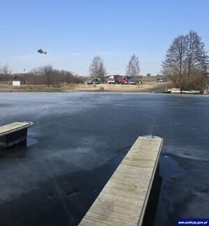 Na brzegu jeziora widoczne samochody służb i lądujący śmigłowiec LPR.