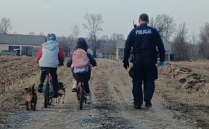 Policjant idzie polną drogą obok dwójki dzieci jadących na rowerach.