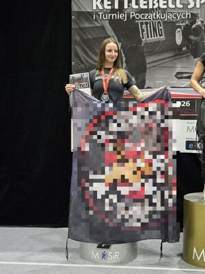 Kobieta na podium, na szyi ma zawieszony medal. W rękach trzyma flagę klubu spotowego.